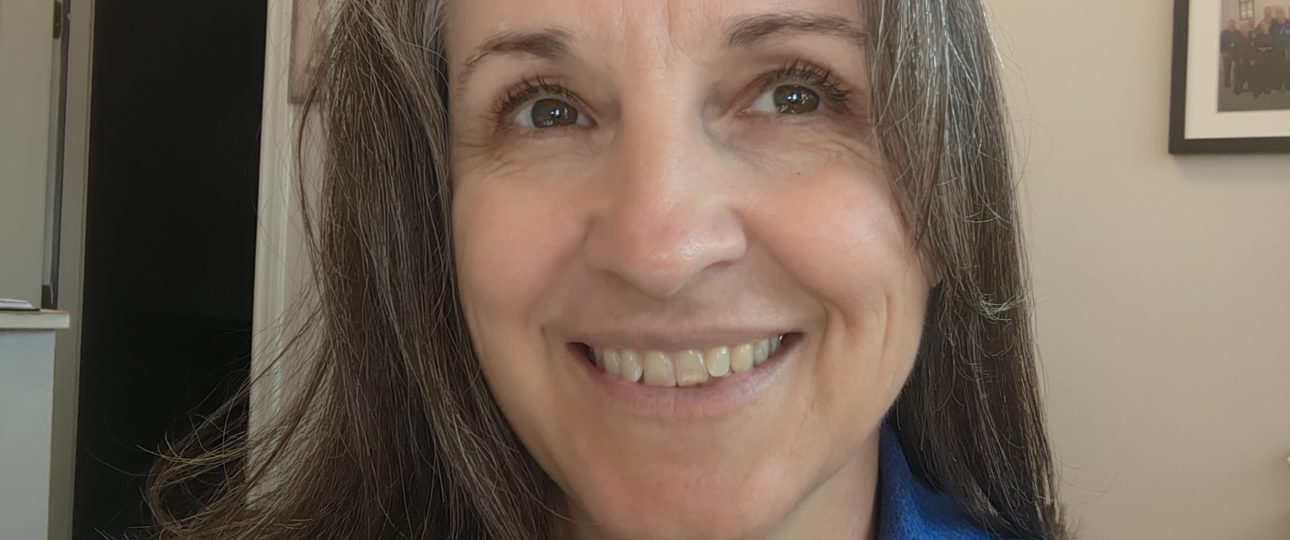Smiling woman with gray hair in a blue polo shirt, indoors with wall art in the background.