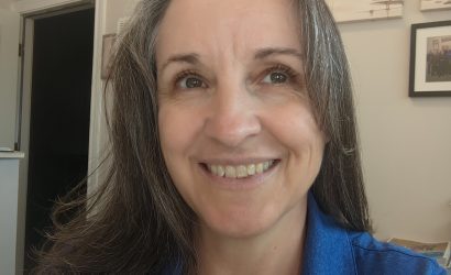 Smiling woman with gray hair in a blue polo shirt, indoors with wall art in the background.